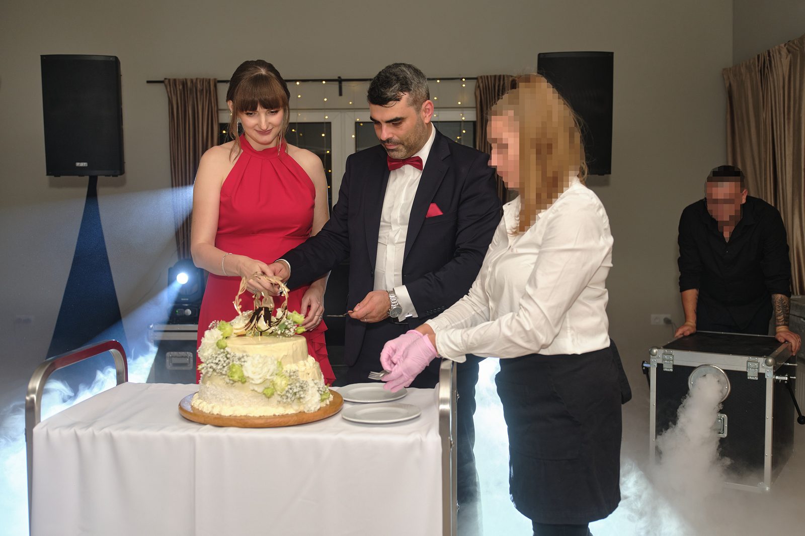 Nowożeńcy stoją przy weselnym torcie; obok znajduje się kobieta w białym stroju.