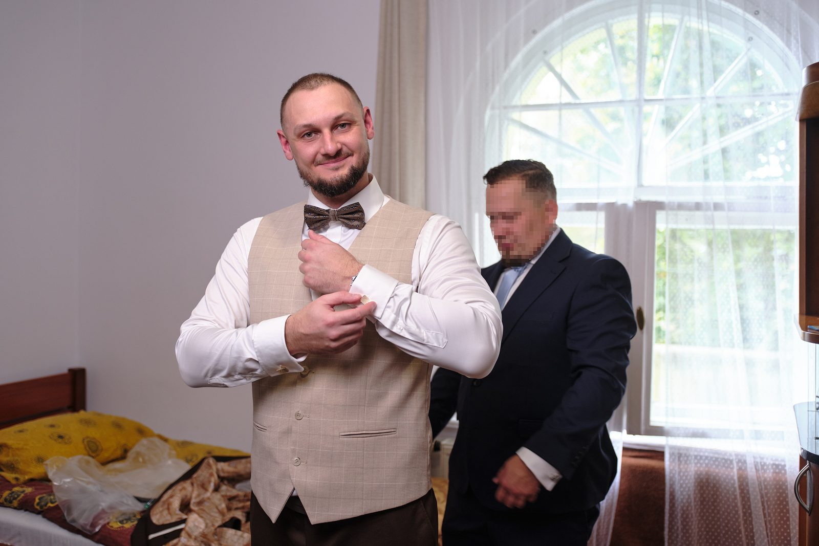 Наречений поправляє краватку-метелик, а позаду стоїть чоловік у темному костюмі.