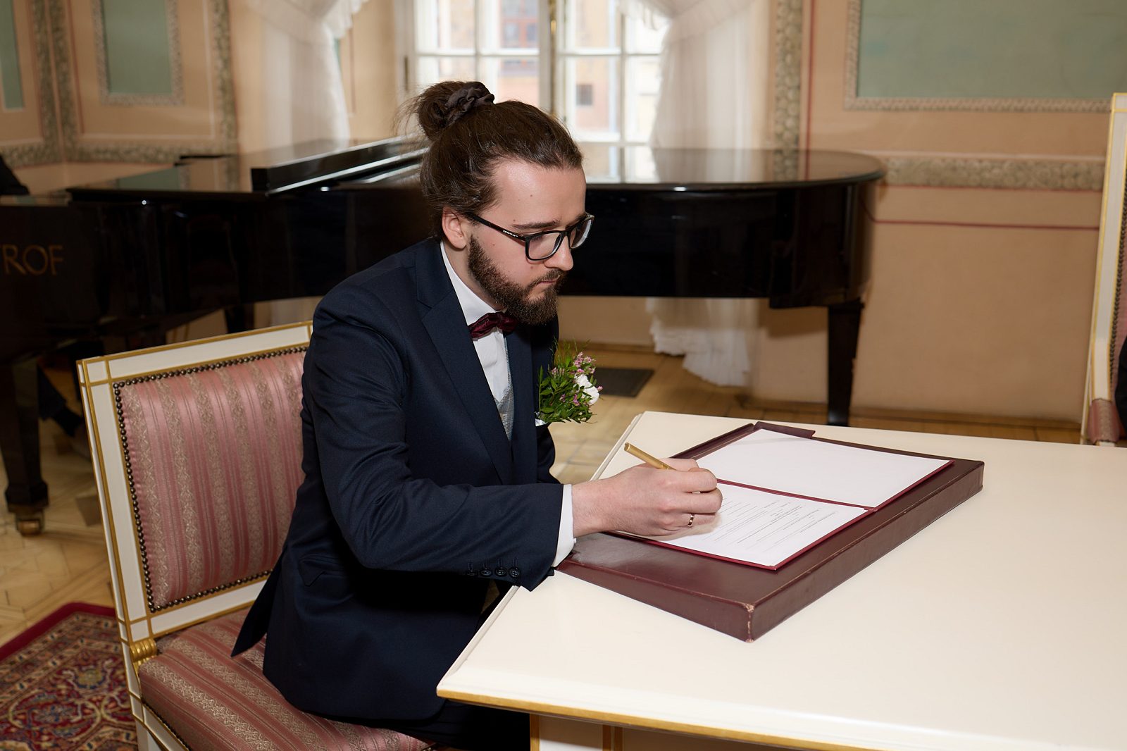 Groom in a navy suit and burgundy bow tie signing the marriage documents