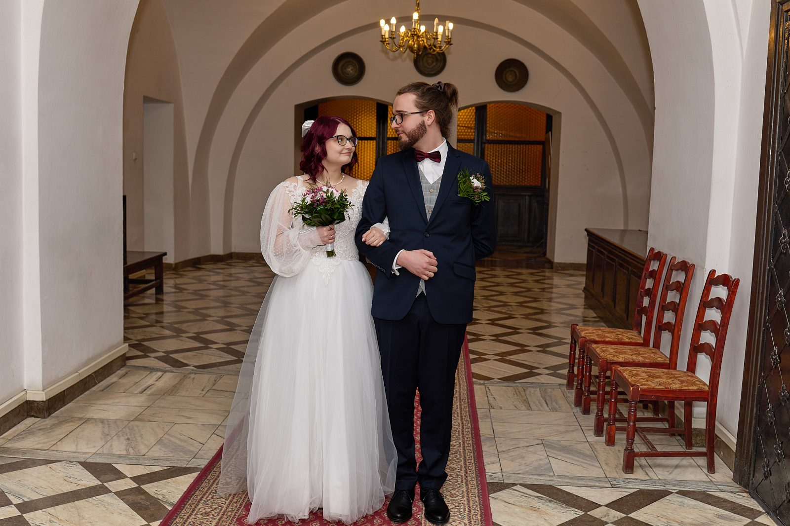 Bride holding a bouquet and linking arms with the groom in a corridor with arched ceilings