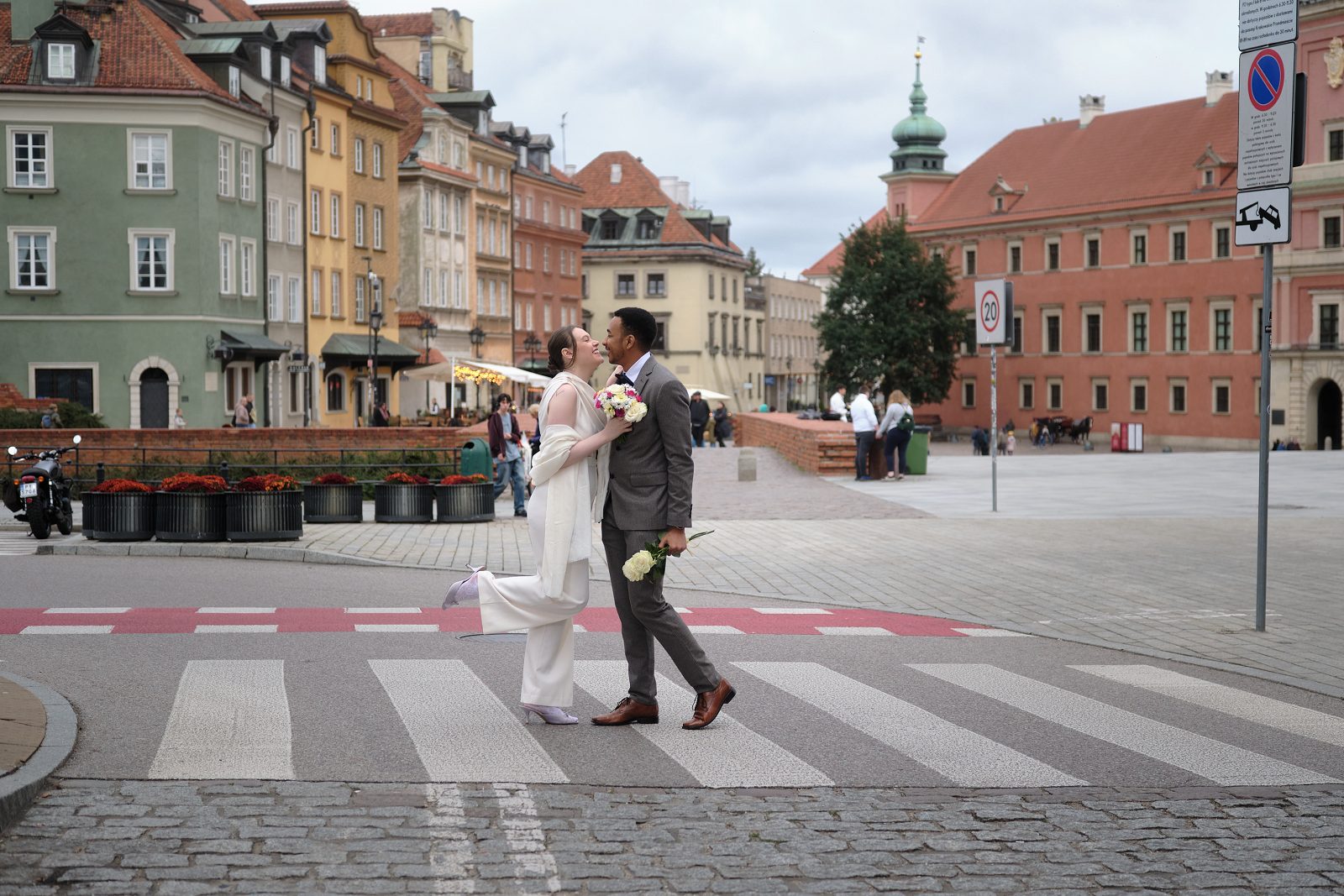 A couple in wedding attire crosses the street with historic Old Town houses behind them.