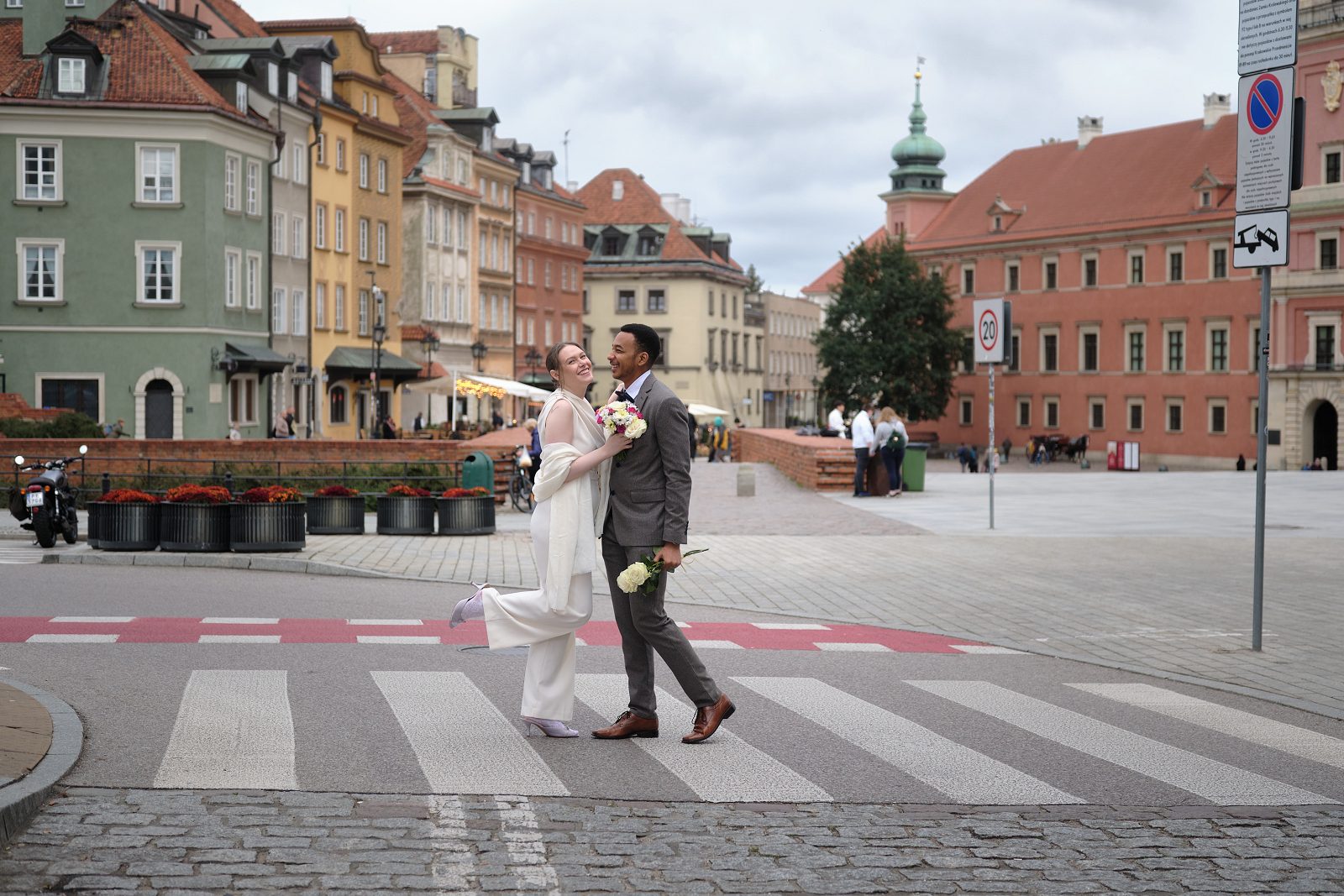 The bride and groom cross a cobbled square among the facades of Warsaw Old Town.