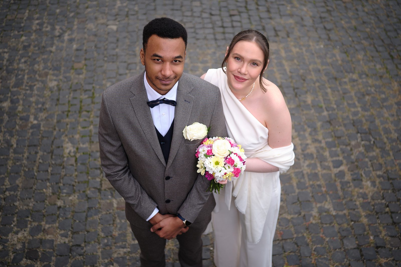 The bride and groom pose by a stone wall as the bride holds her bouquet.