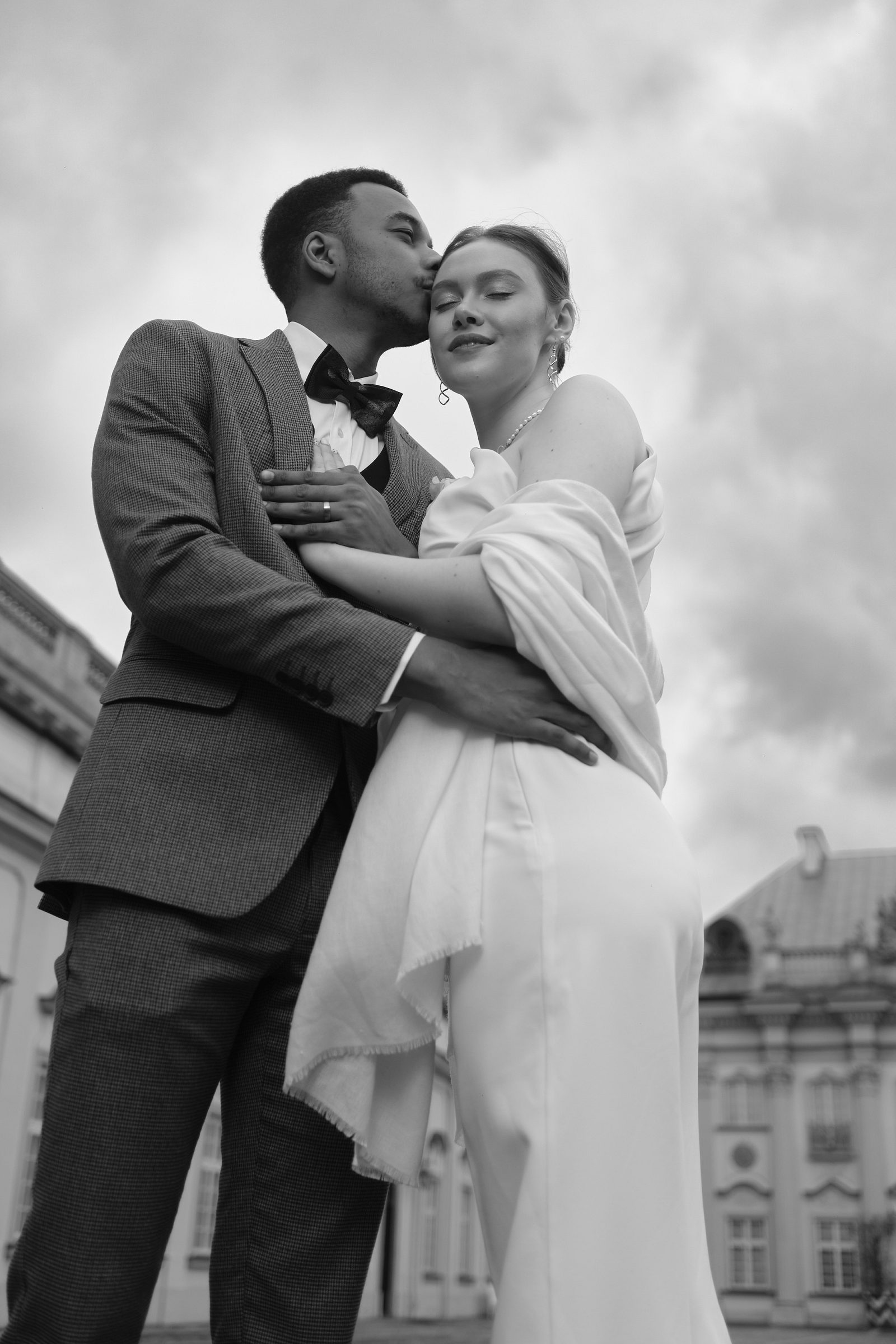 The bride and groom embrace in front of a historic stone facade.