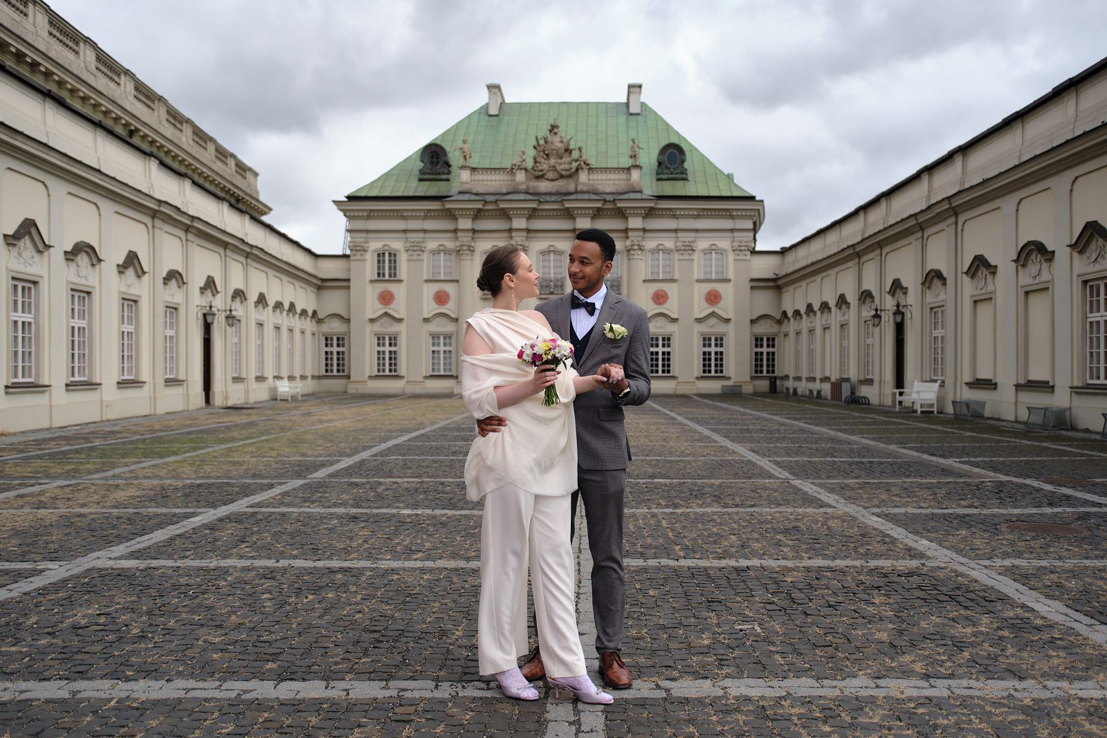 The bride and groom stand in a cobbled courtyard before a historic building.