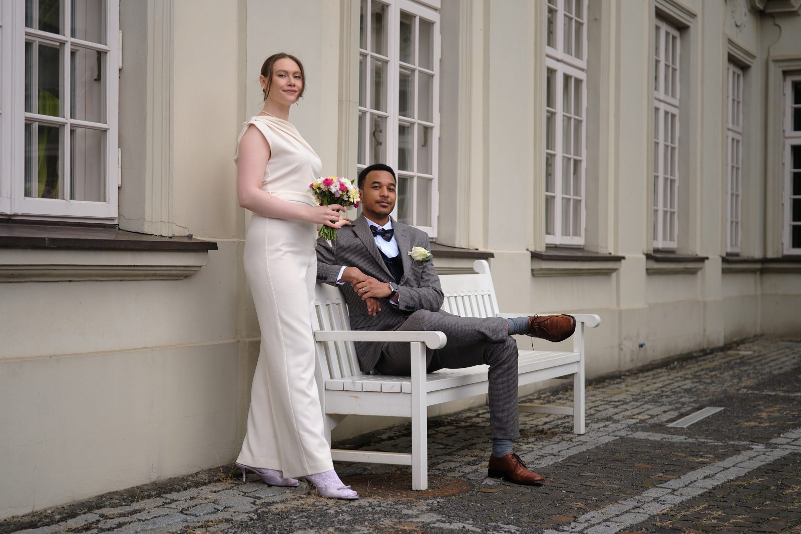 The bride stands with her bouquet beside a white bench where the groom is seated.