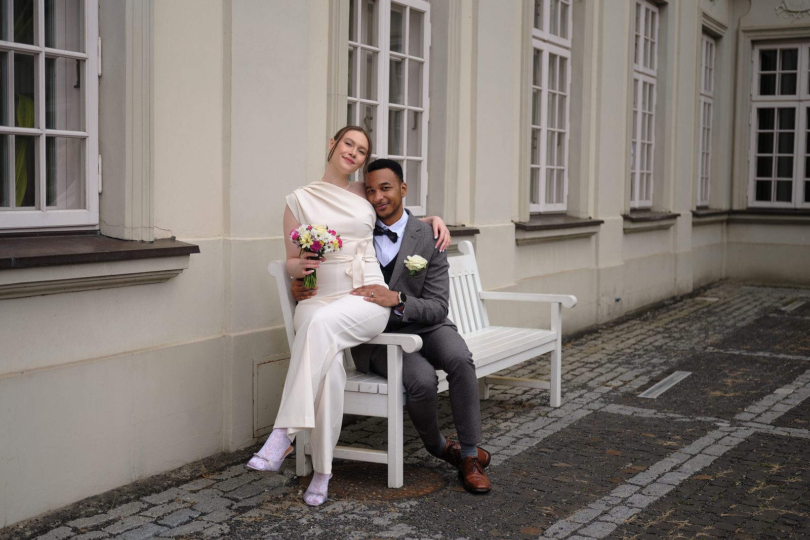 The bride and groom sit together on a white bench beside tall windows.