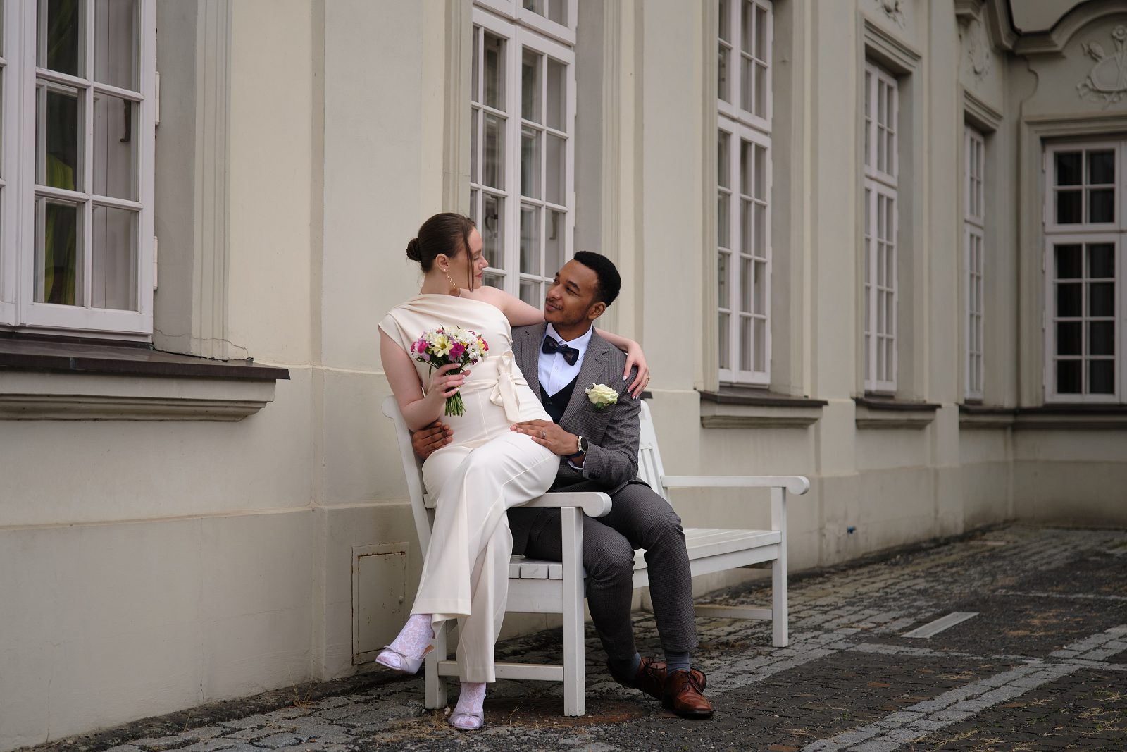 The couple sit on a bench by a light facade as the bride turns toward the groom.