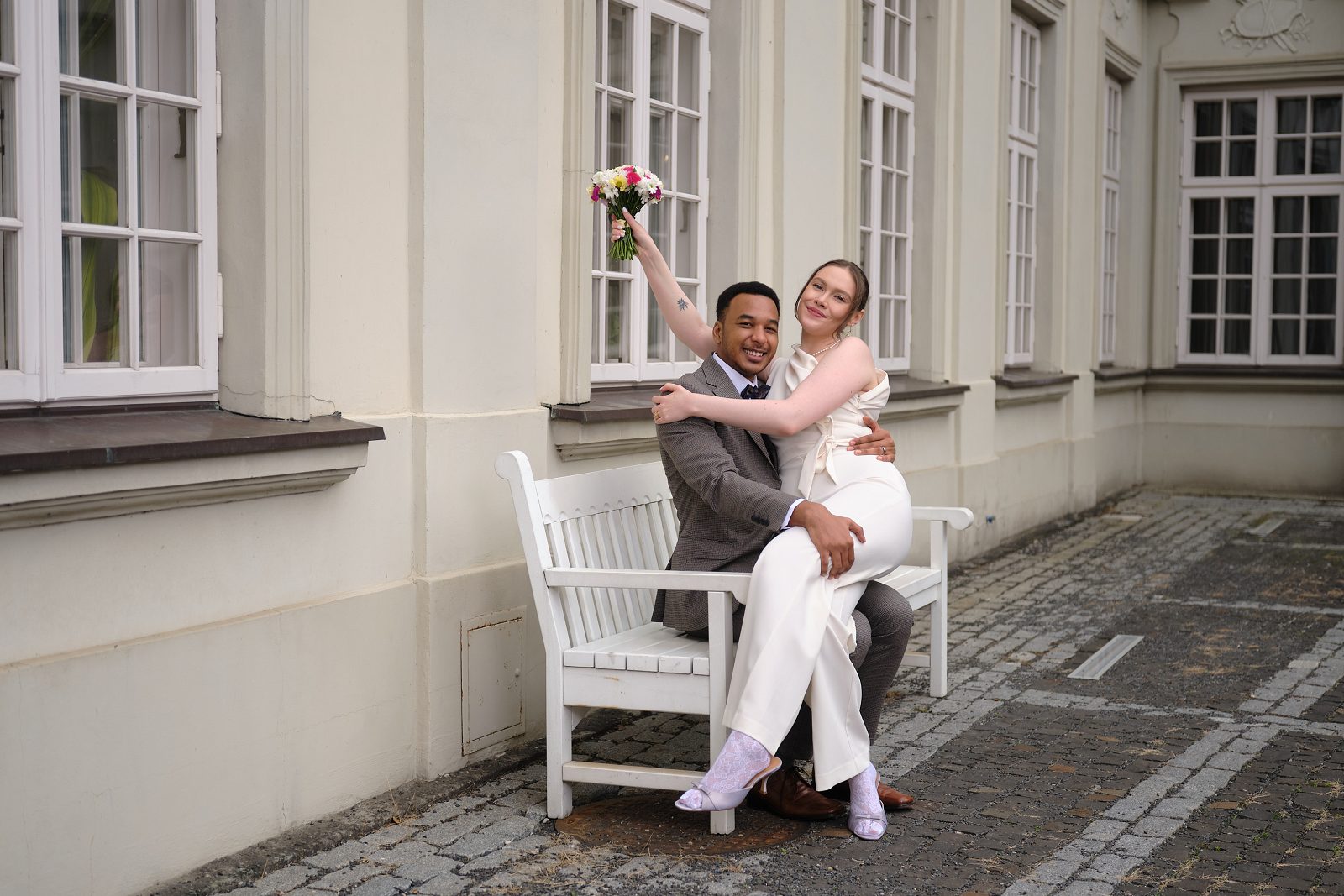 The bride lifts her bouquet above the seated groom beside a white bench.
