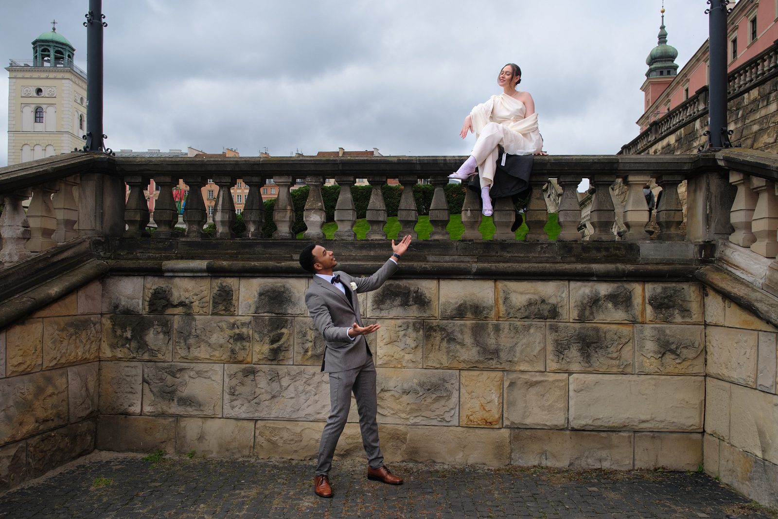 The bride sits on a stone balustrade above the groom standing beside the wall.
