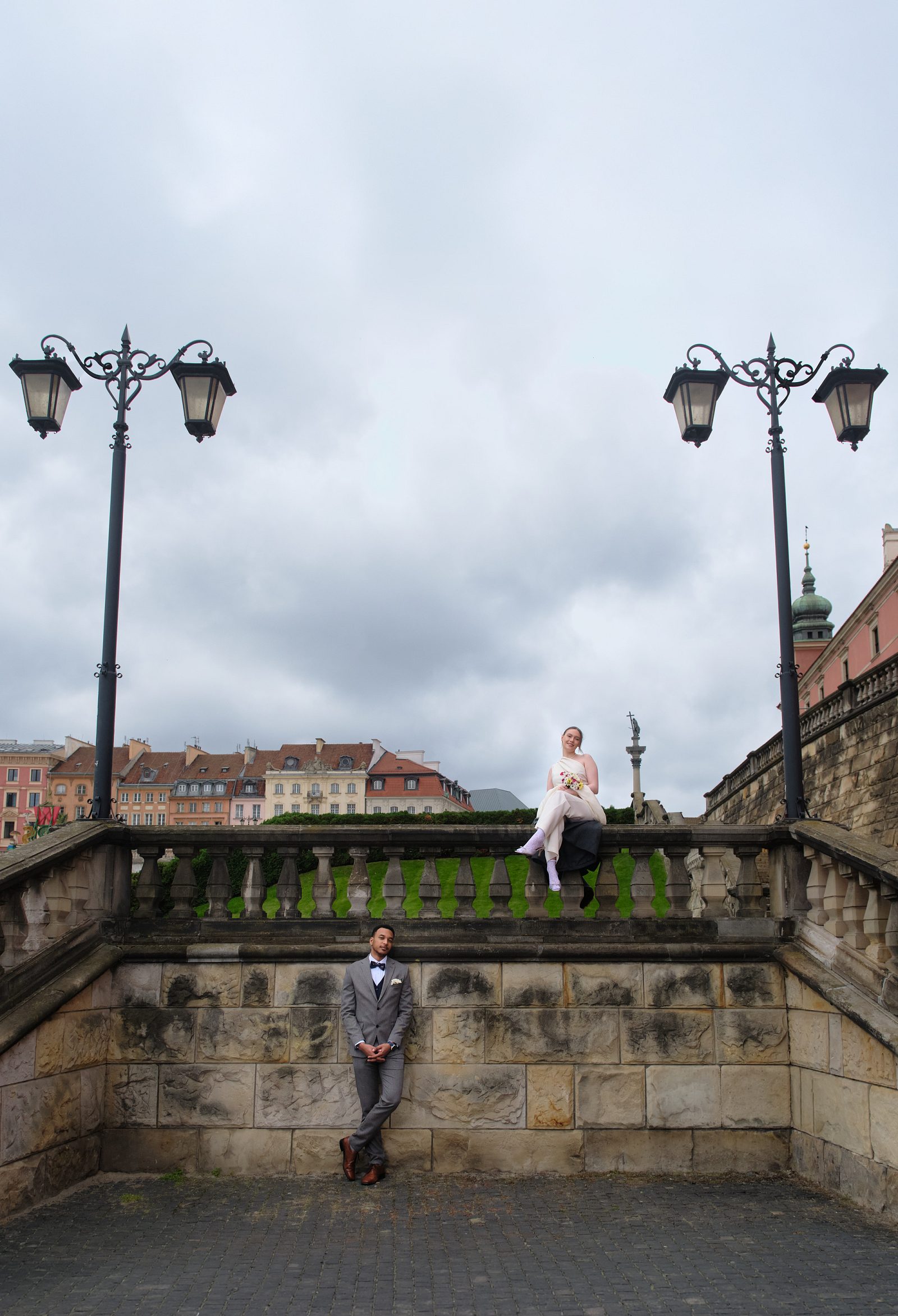 The bride sits on a stone parapet while the groom stands below beside the wall.