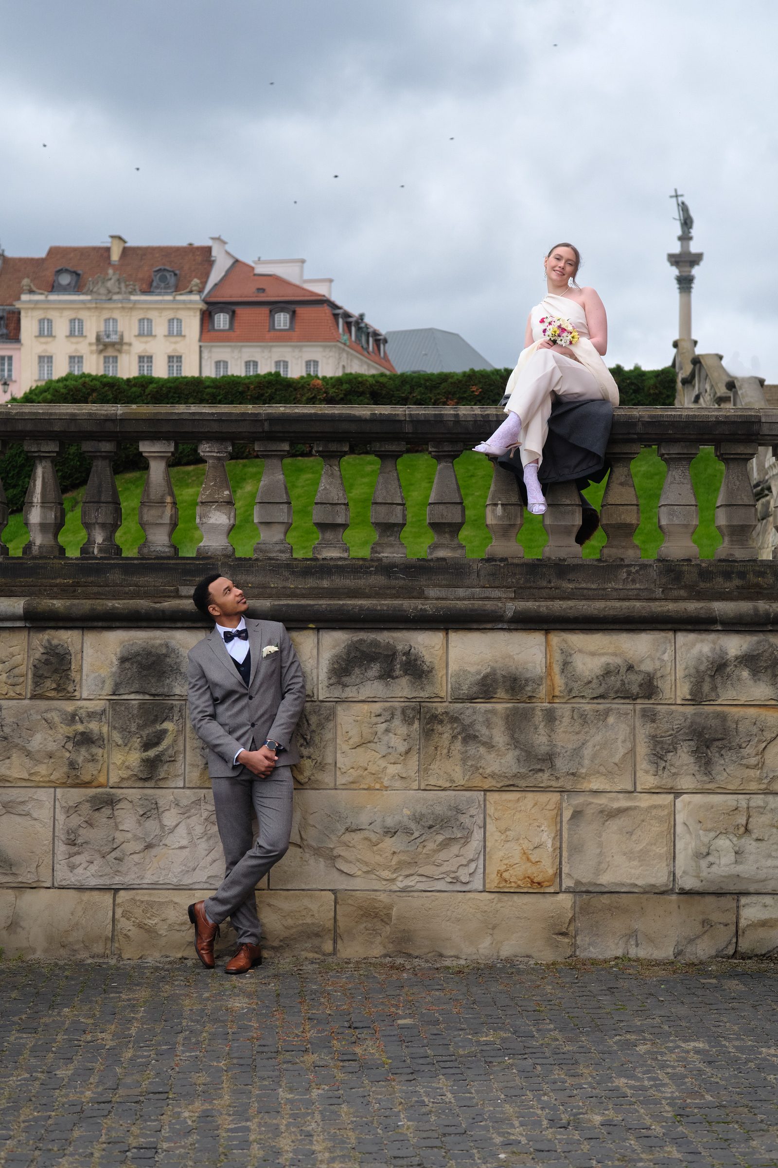 The bride sits on a stone parapet above the stairs while the groom stands below.