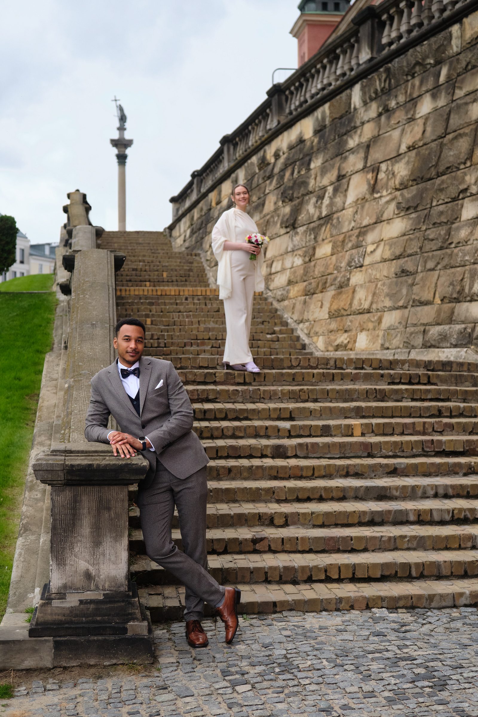The bride stands with her bouquet by a stone staircase while the groom sits on nearby steps.