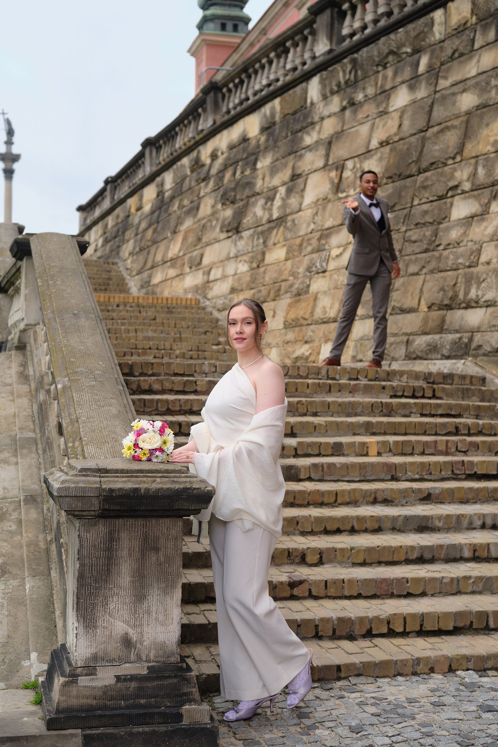 The bride poses beside a stone pedestal near an old staircase.