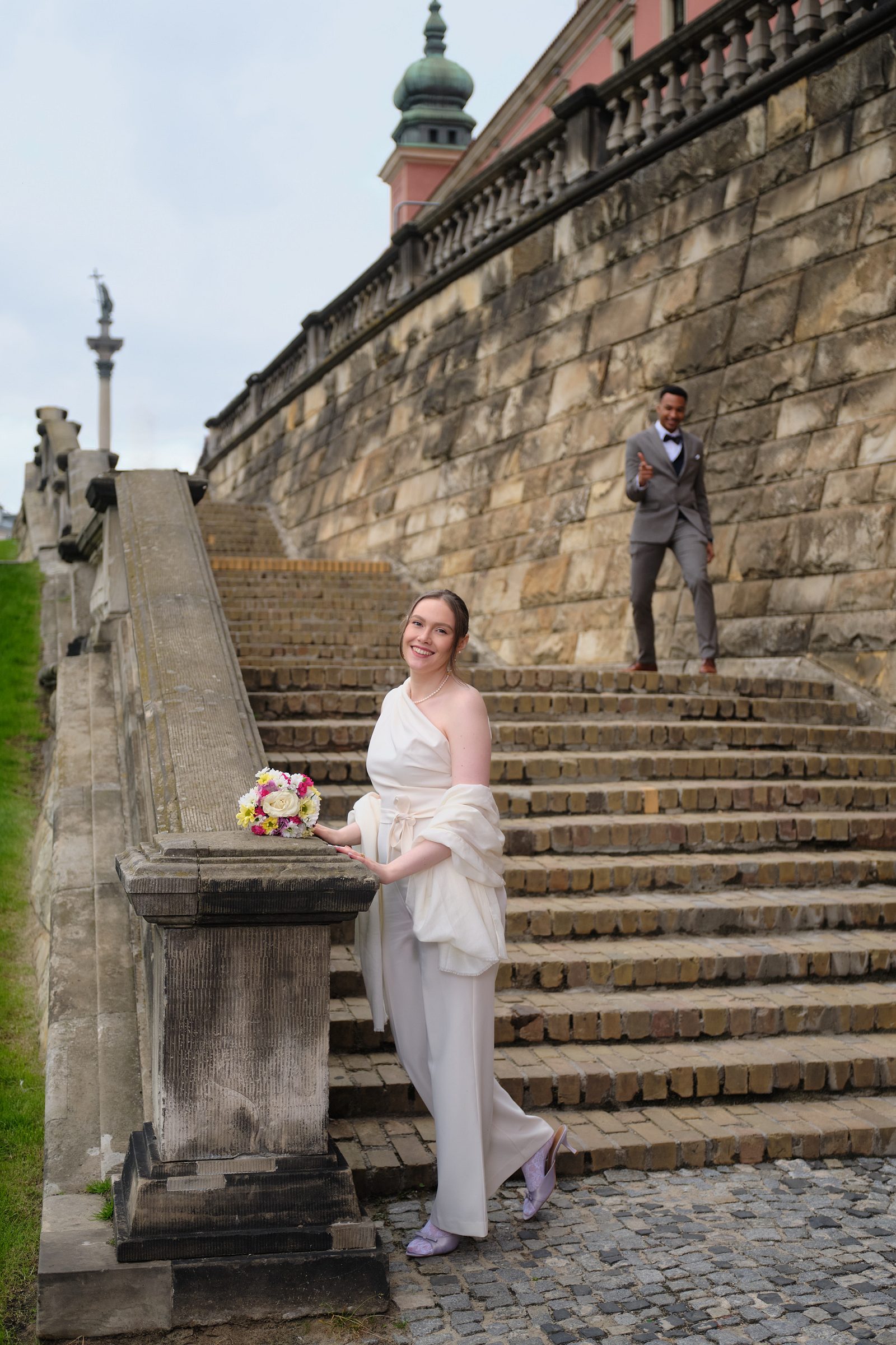 The bride stands beside a stone railing with a staircase rising behind it.