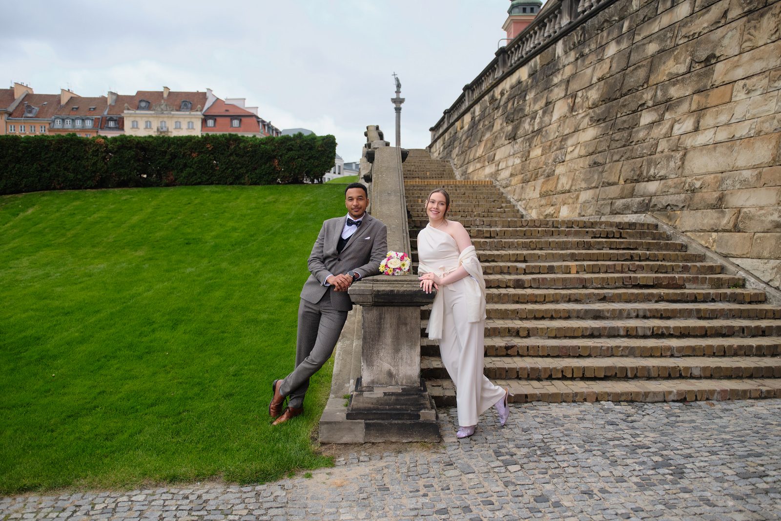 The bride and groom sit on a stone parapet by the stairs with a bouquet between them.