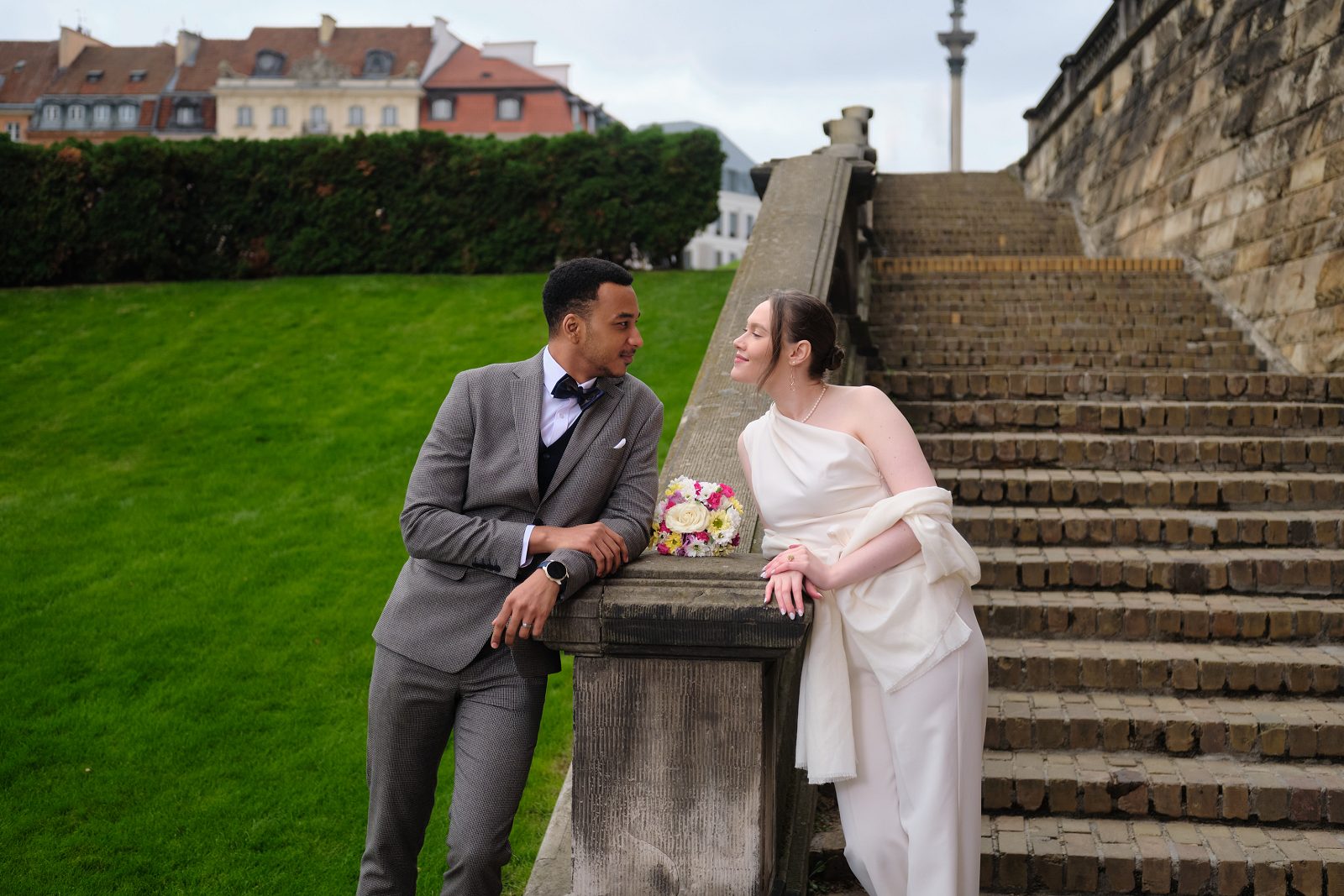 The bride sits with her bouquet on a stone parapet beside the groom.