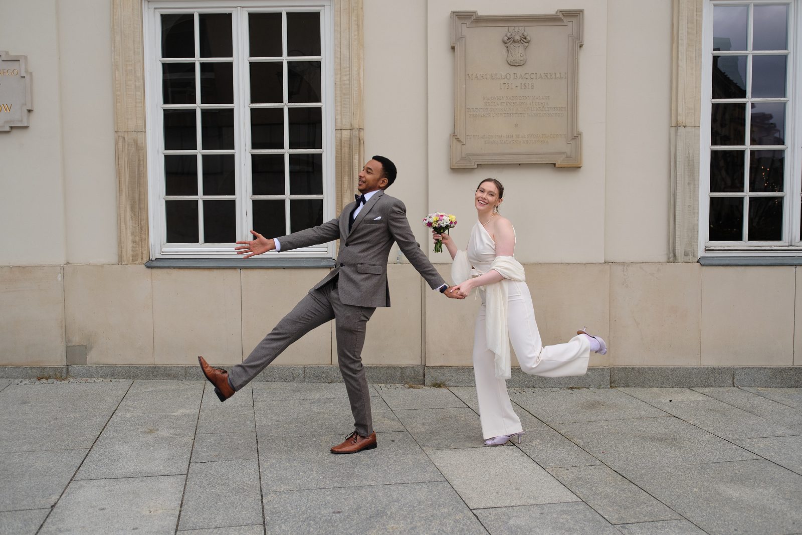 The bride and groom pose playfully in motion on a cobbled square by an old facade.