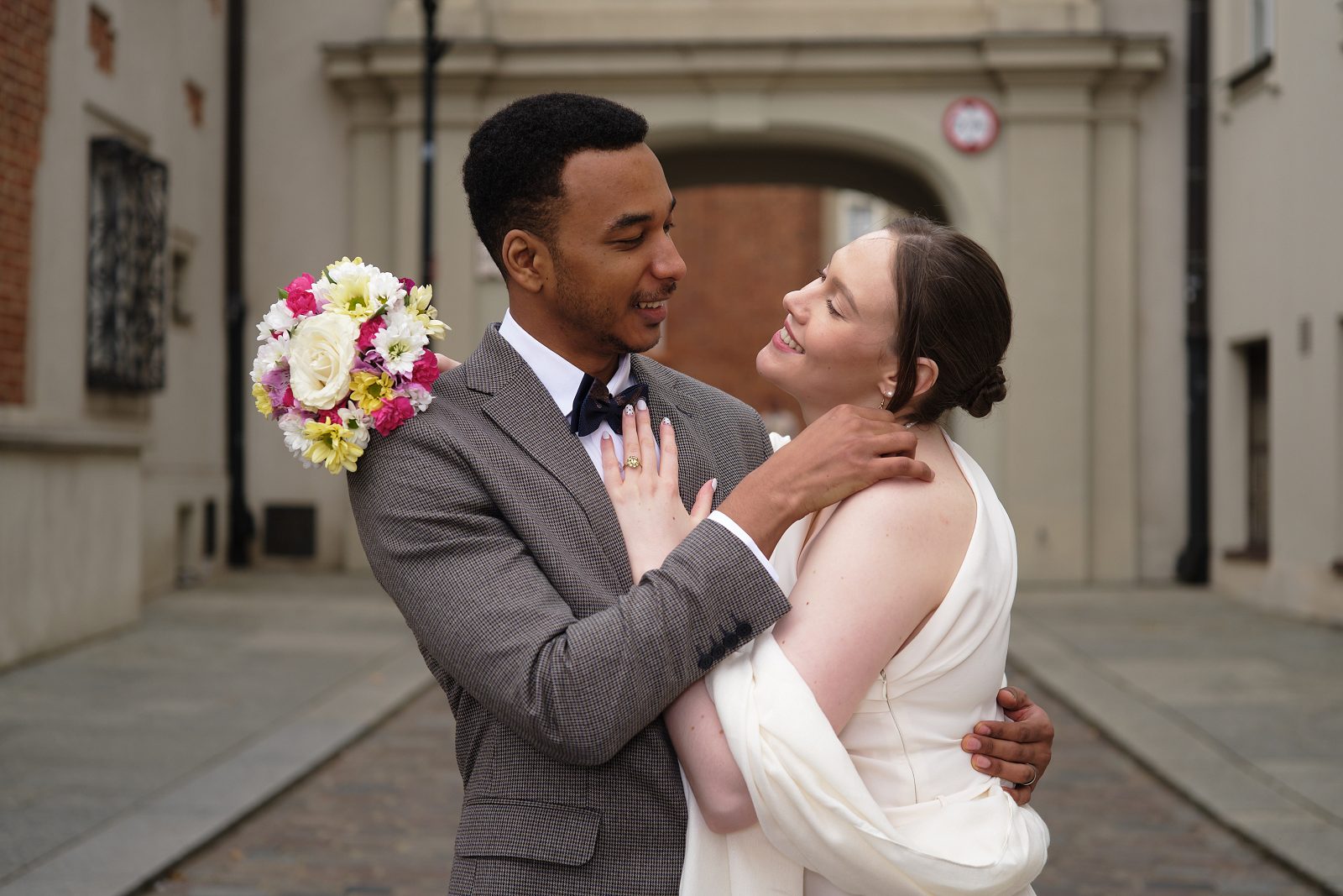 The bride holds her bouquet by her shoulder as the groom embraces her beneath a historic arch.