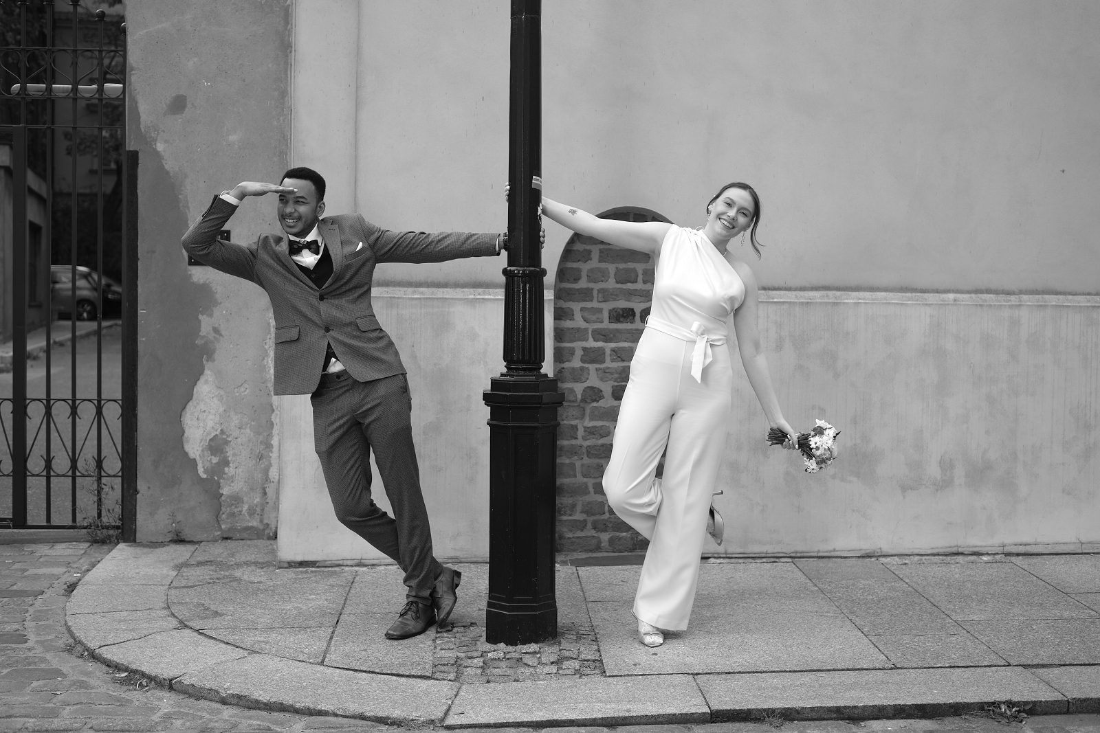 Black-and-white frame of the couple posing by a street lamp with their arms extended.