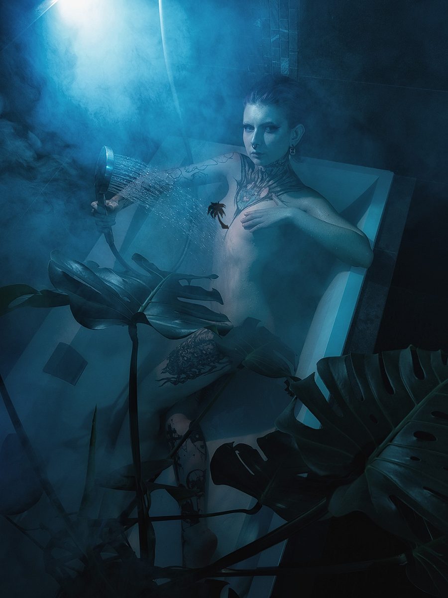 Tattooed woman in a bath among large leaves and blue mist
