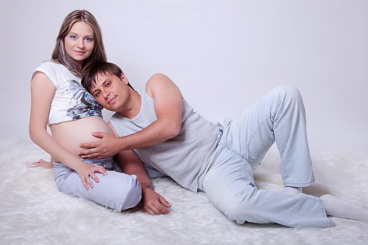 Мужчина лежит рядом с беременной женщиной на белом покрывале