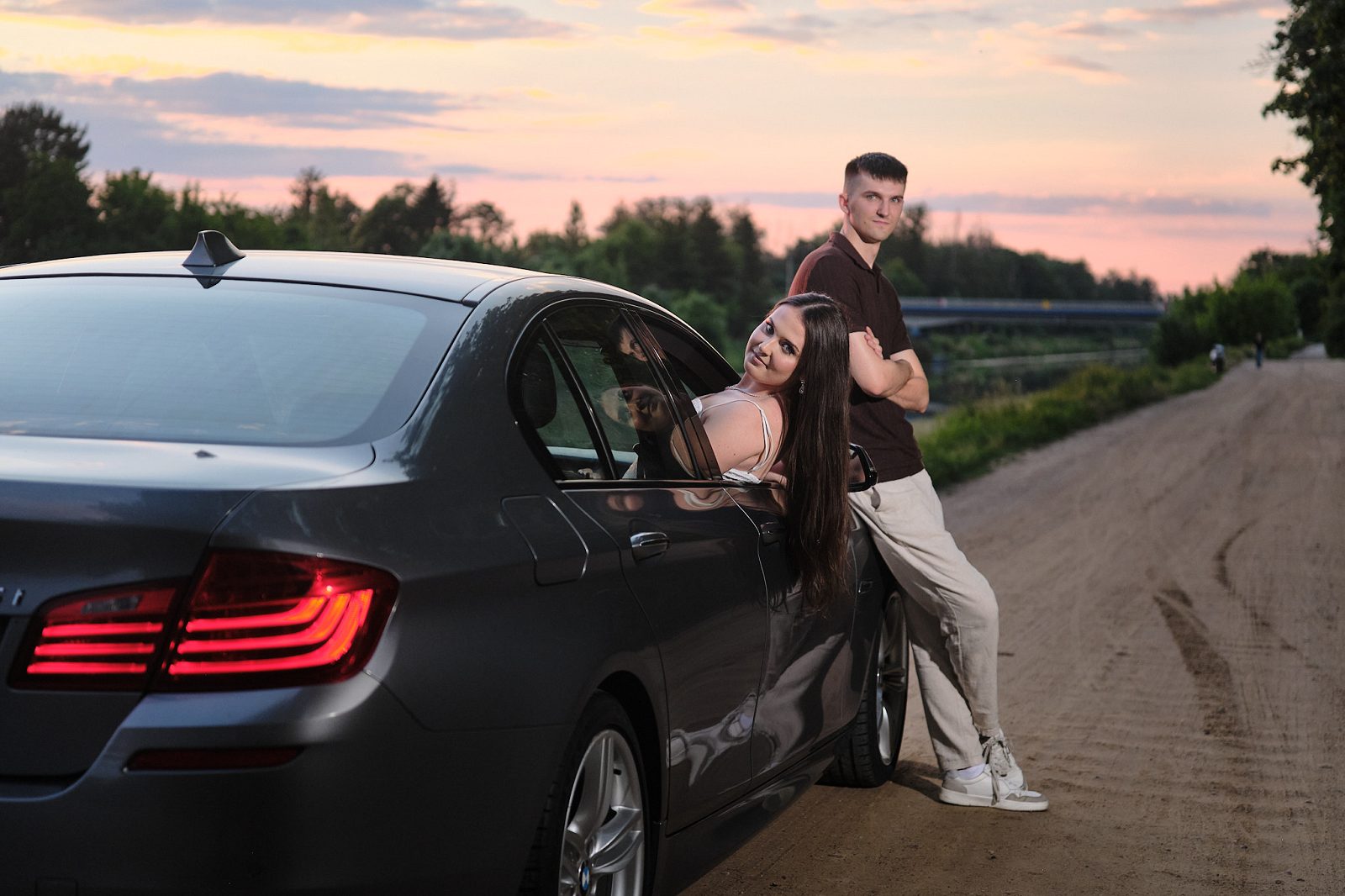 Couple beside a car on a dirt road at sunset; the woman looks out of the window