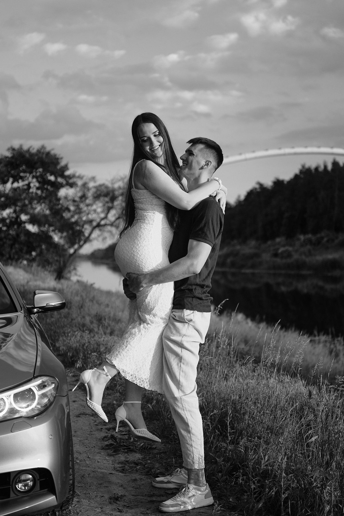 Black-and-white portrait of a couple embracing beside a car by the water