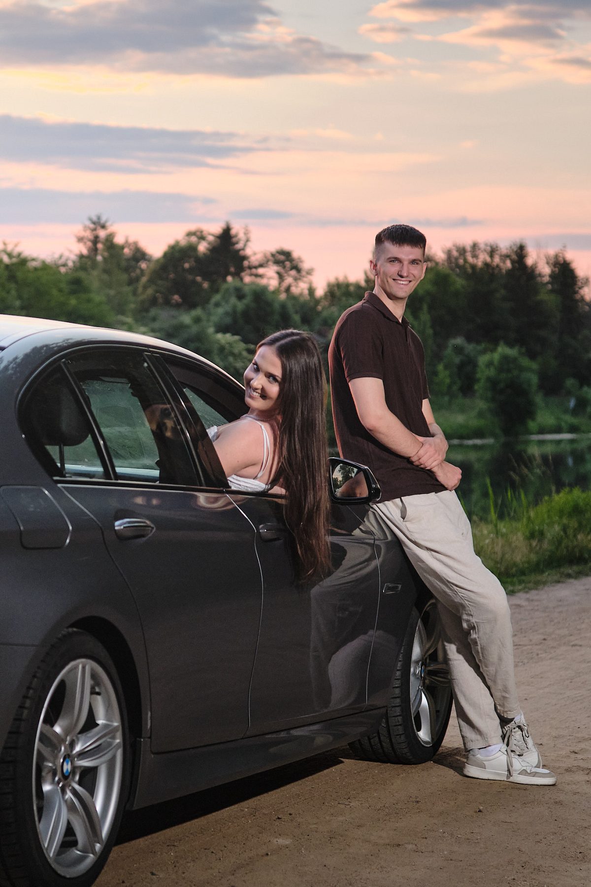 Man leaning against a car while a woman looks out of the window under the evening sky