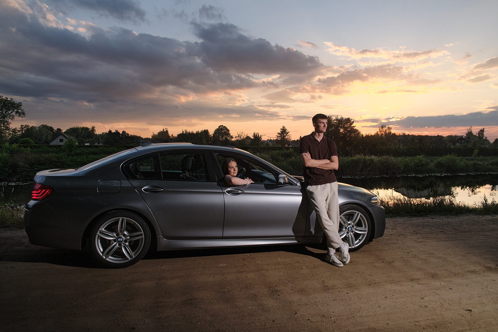 Man standing beside a car while a woman sits inside by the water at sunset