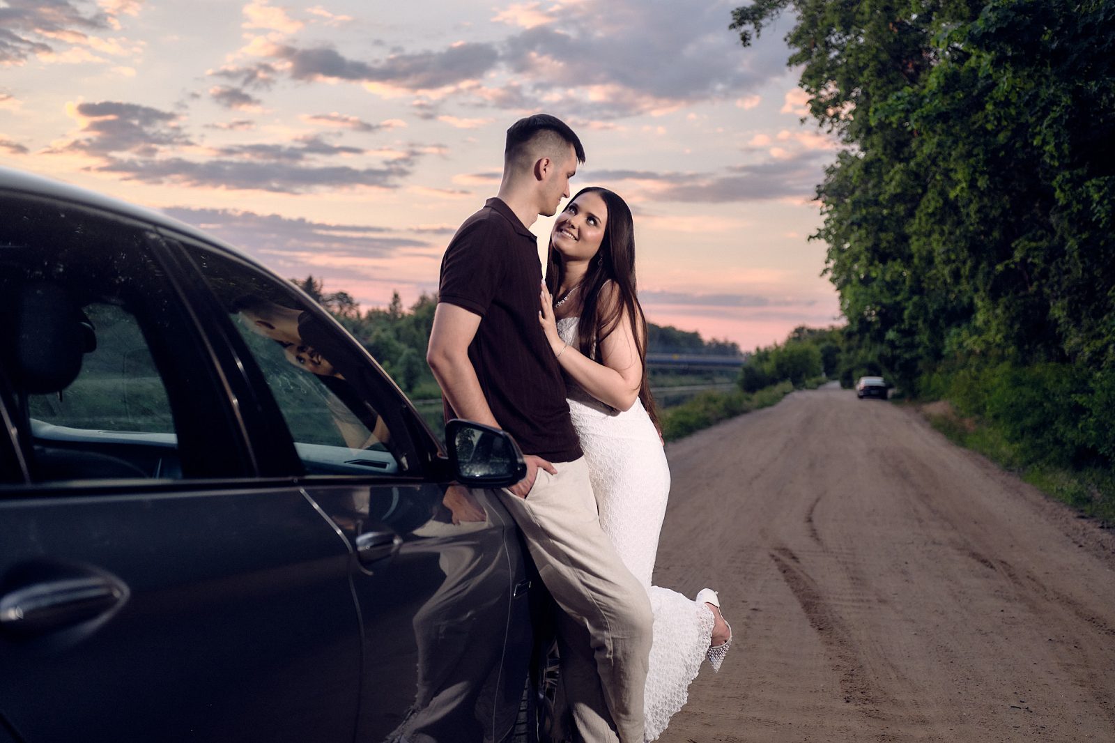 Couple embracing beside a car on a dirt road at sunset