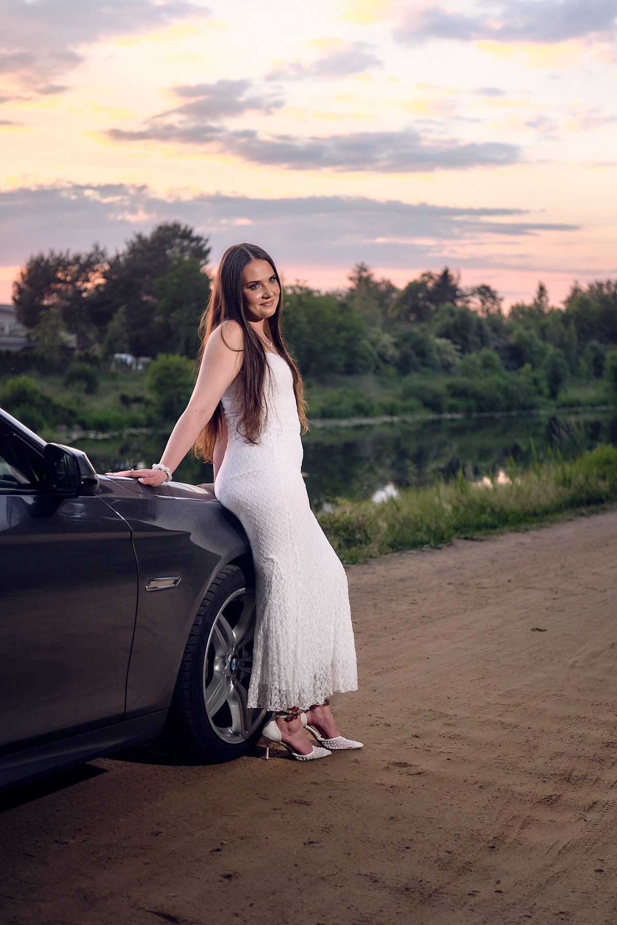 Woman in a white dress leaning against a car under the evening sky