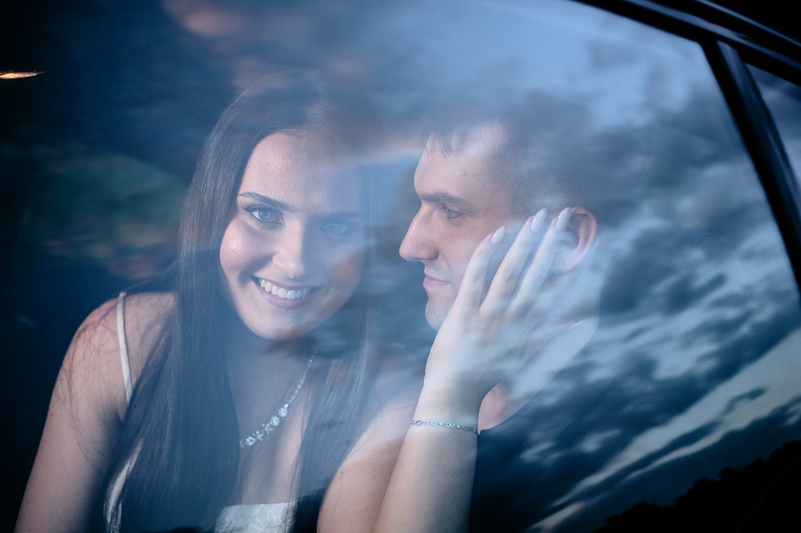 Black-and-white portrait of a couple in a car photographed through glass, with the man touching the woman’s face