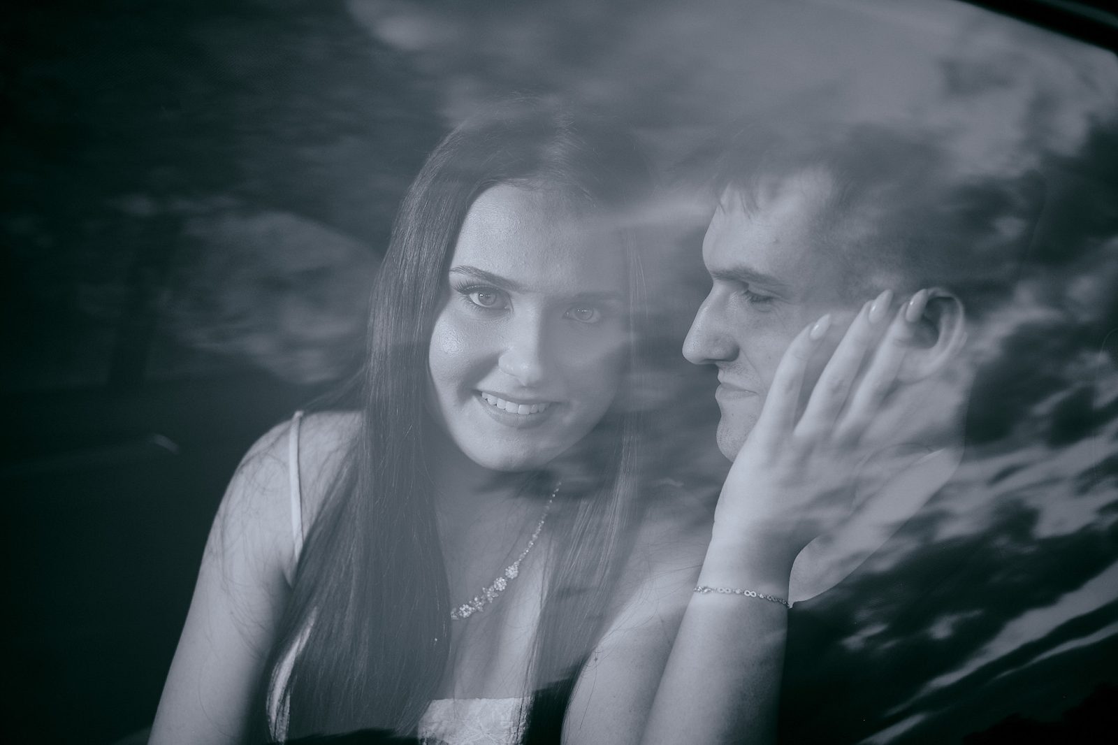 Black-and-white portrait of a couple photographed through a car window, with the man touching the woman’s face