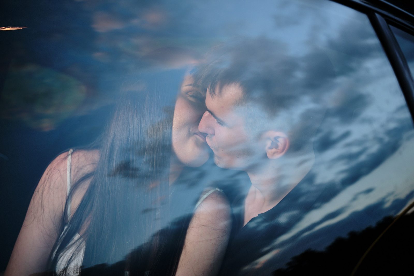 Couple kissing inside a car, photographed through glass reflecting the sky