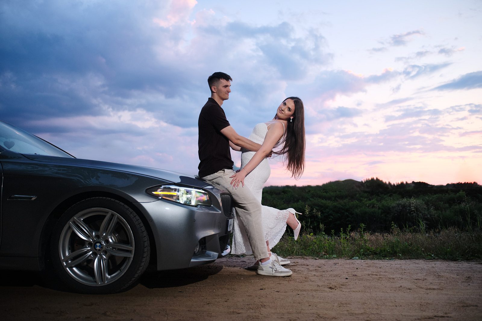 Man sitting on a car hood while a woman leans against him under the evening sky