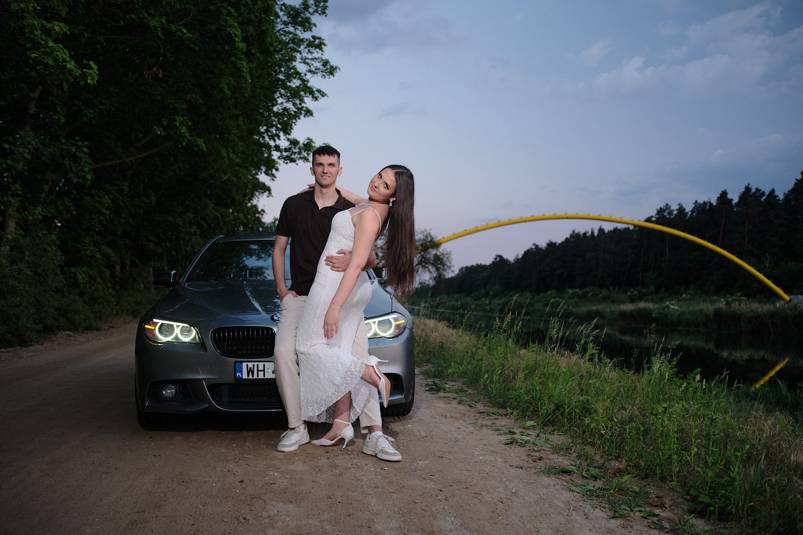 Couple standing in front of a car by the water with a yellow arched bridge behind them
