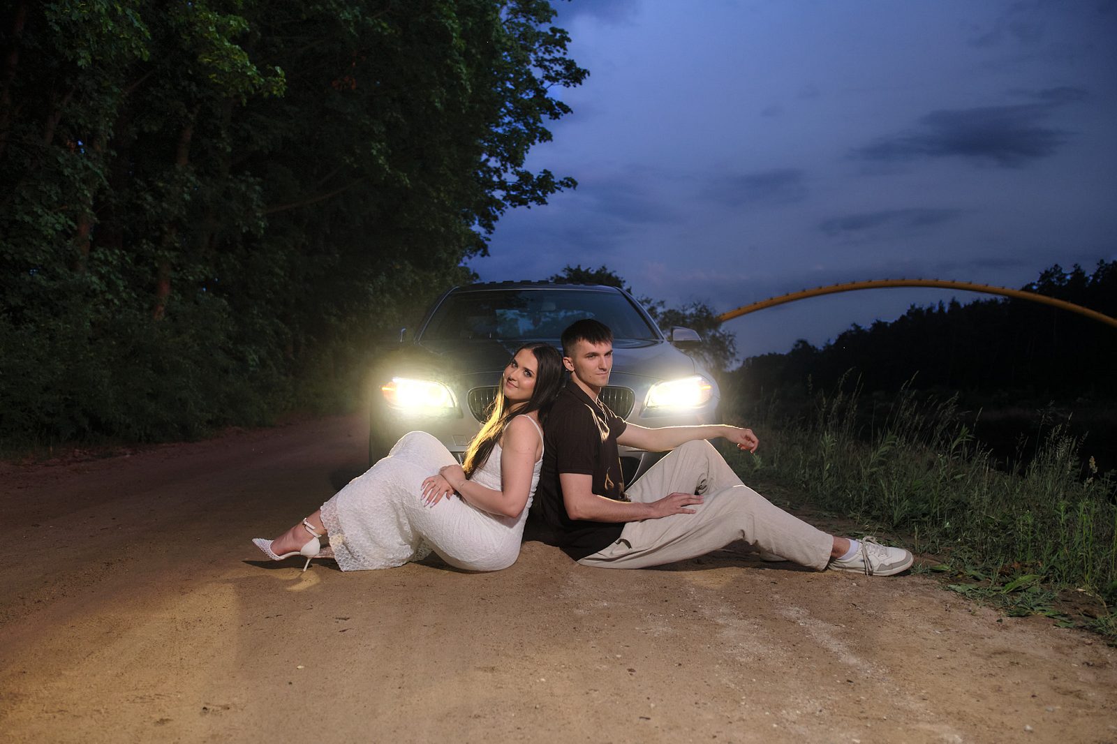 Couple sitting in front of a car with its headlights on at dusk