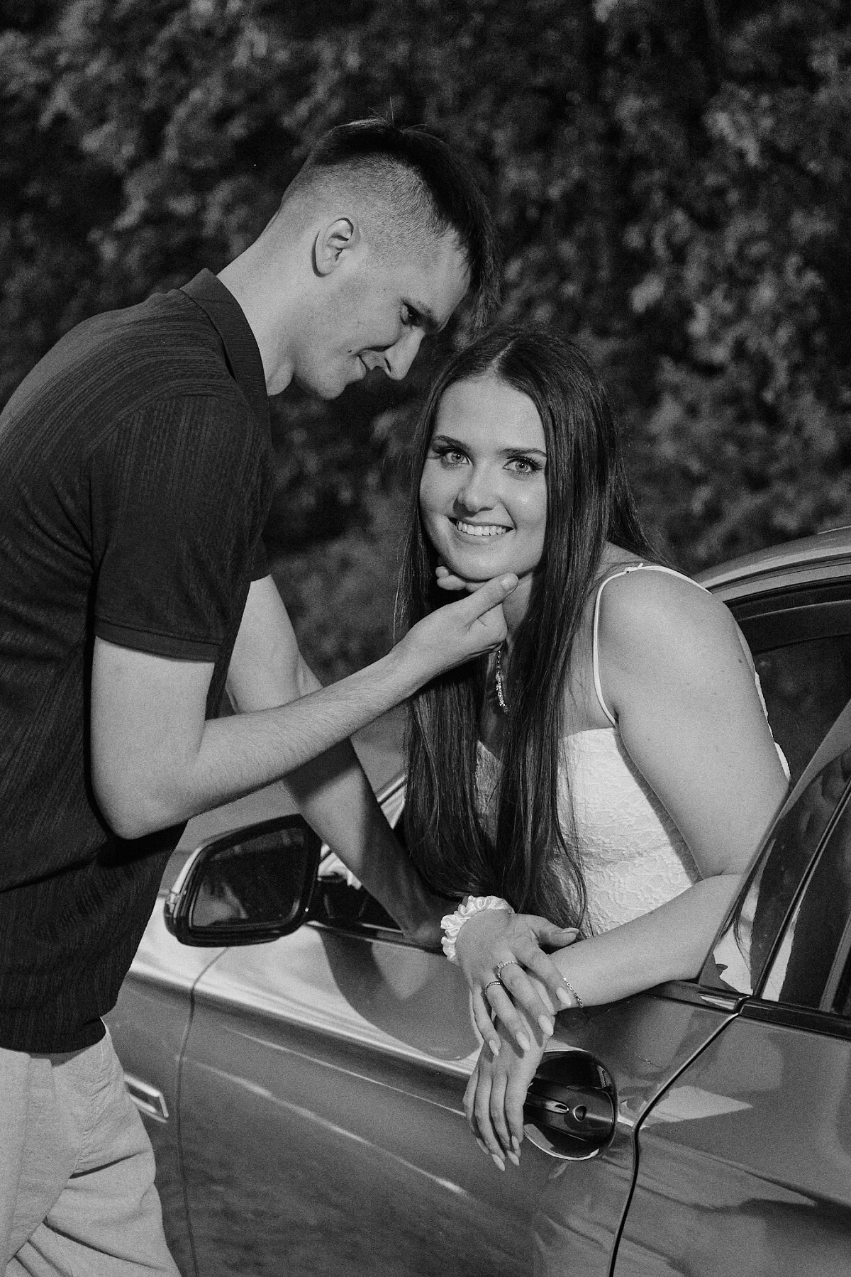 Black-and-white portrait of a couple beside a car, with the man touching the woman’s chin