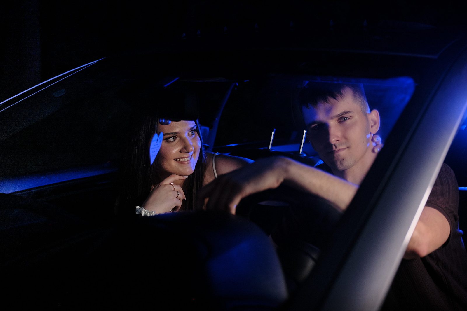 Couple inside a car in blue night lighting; the man is behind the wheel