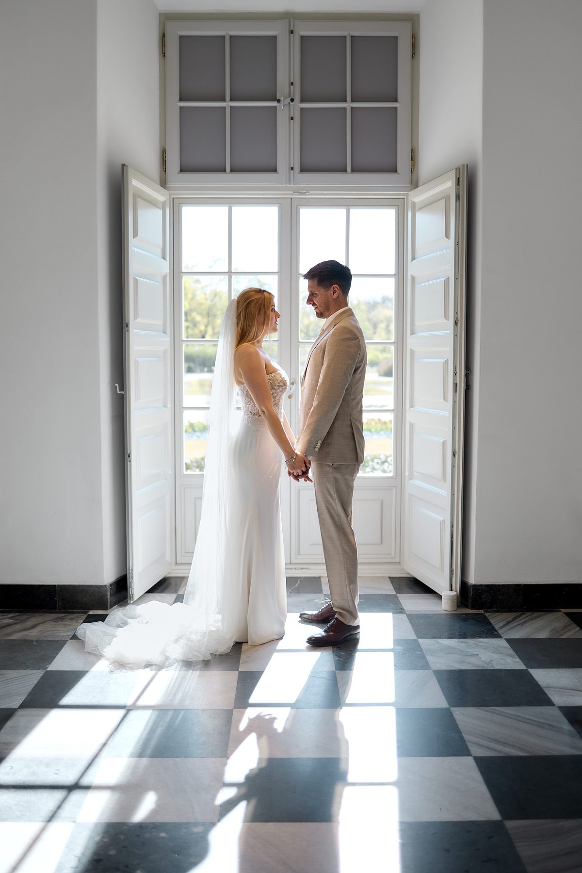 The newlyweds hold hands in the doorway of a bright grand interior with a black-and-white marble floor.