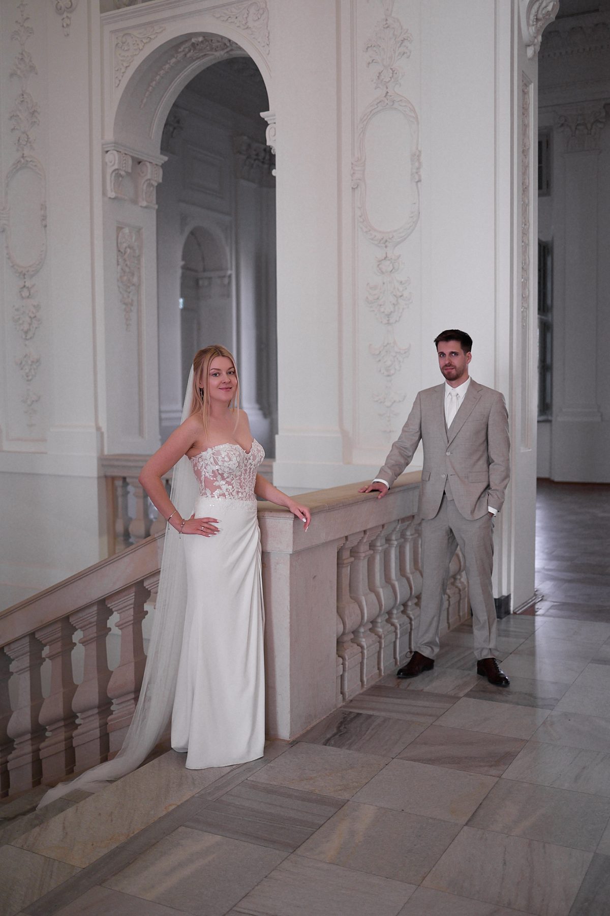 The newlyweds hold hands beside a balustrade in a spacious bright hall.