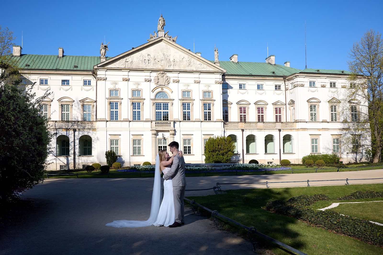 The newlyweds kiss in front of the light-colored palace beneath a blue sky.