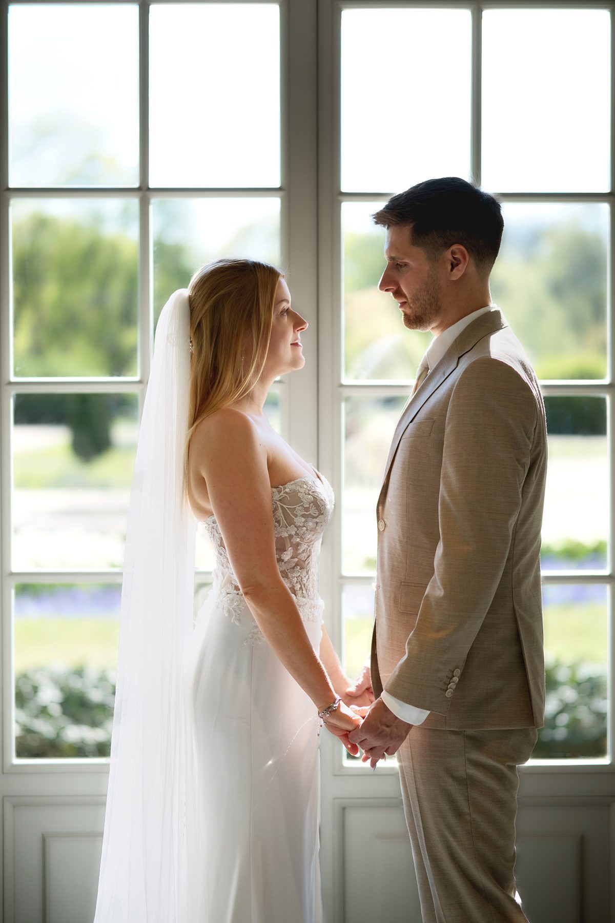 The bride and groom hold hands by tall windows and look at each other.