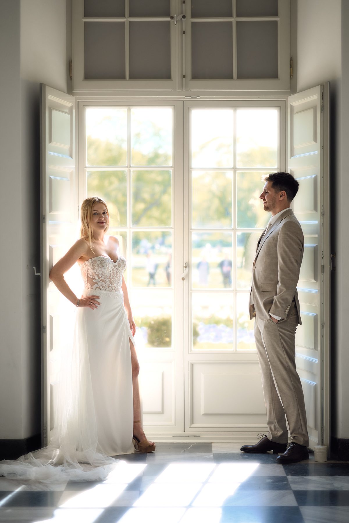 The bride stands by open doors while the groom looks at her in a bright hall with a marble floor.