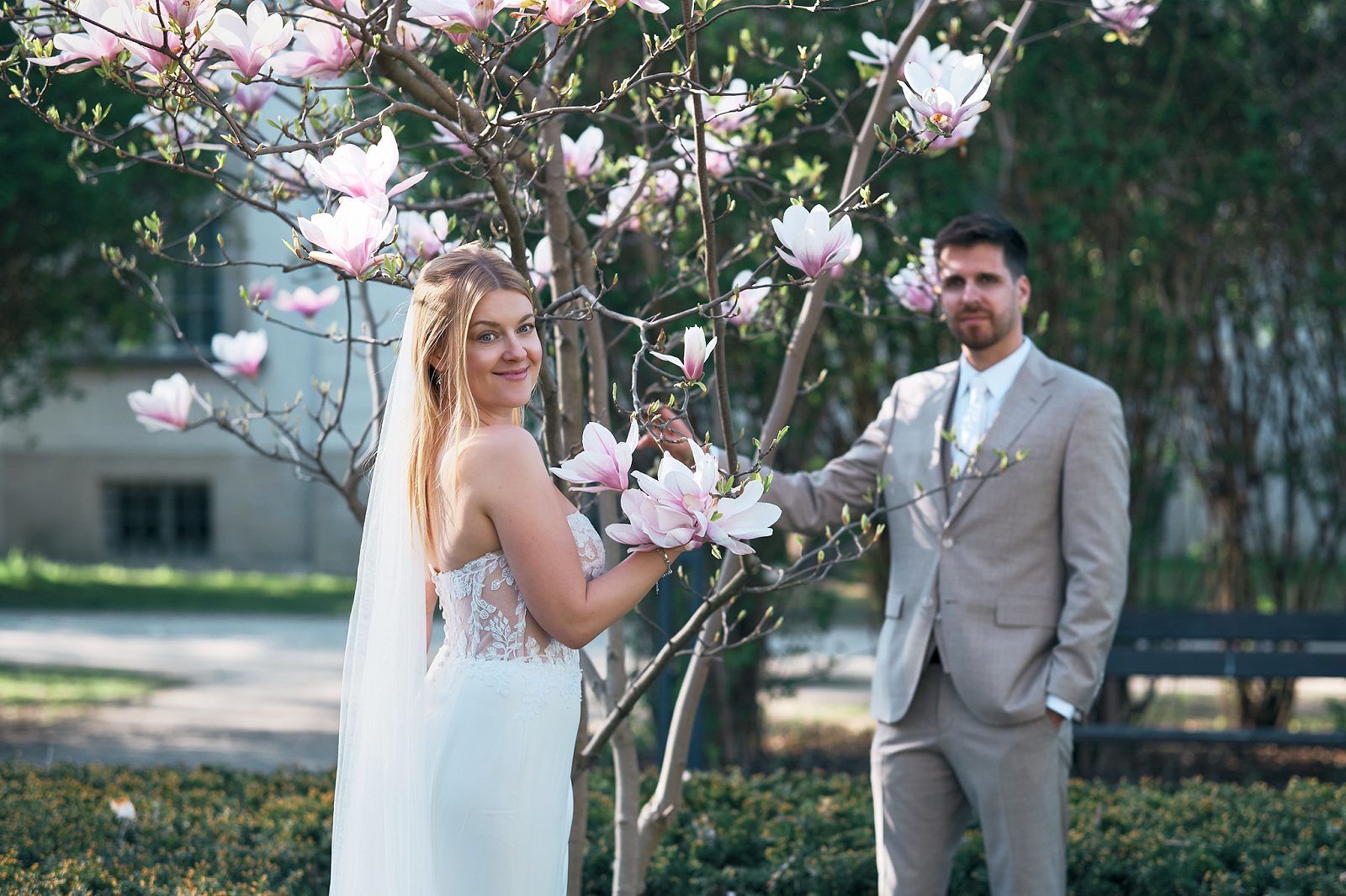 The bride holds a blooming magnolia branch while the groom stands behind her in the garden.