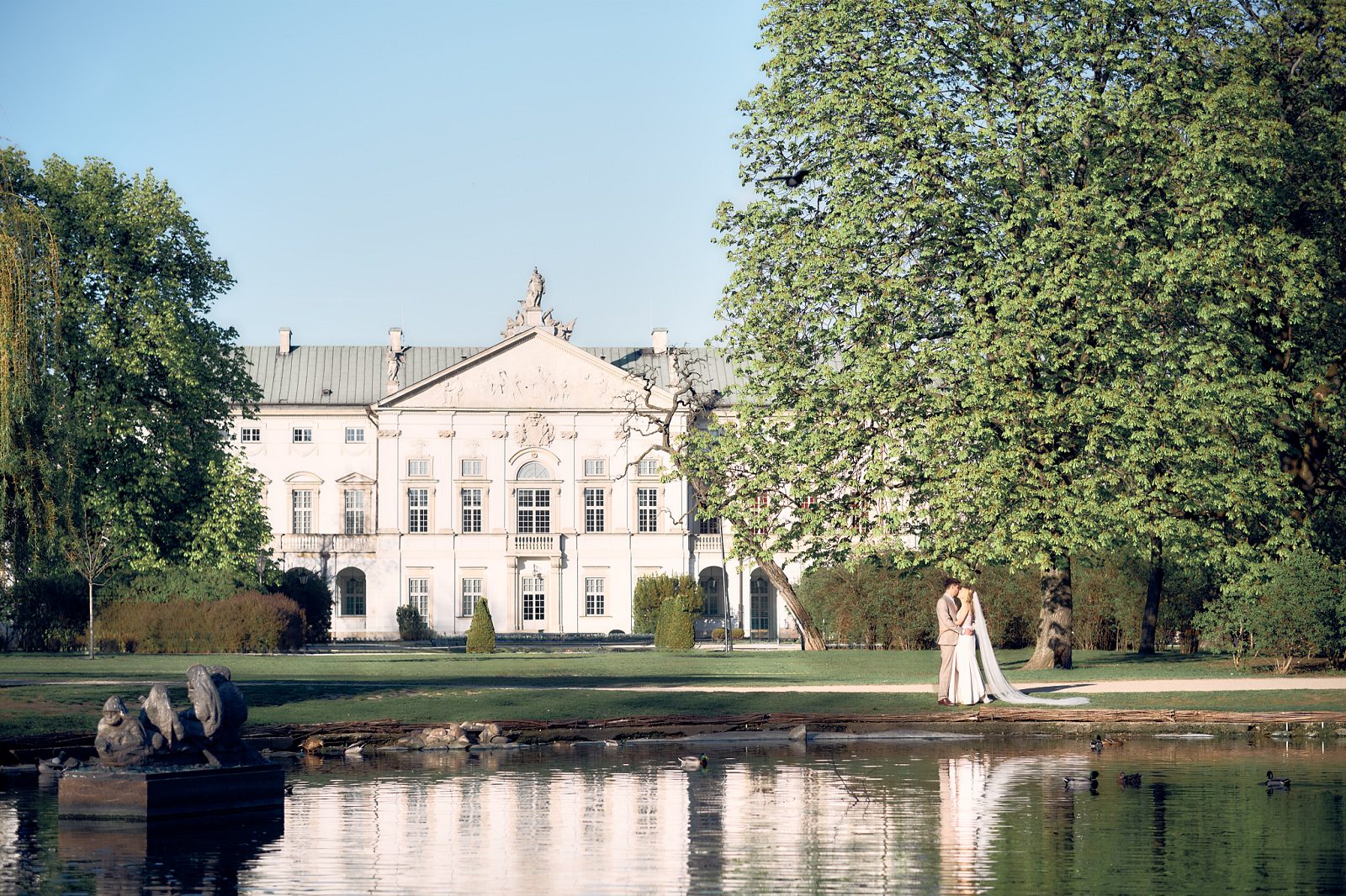 The newlyweds stand beside a pond in the garden with the light-colored palace behind them.