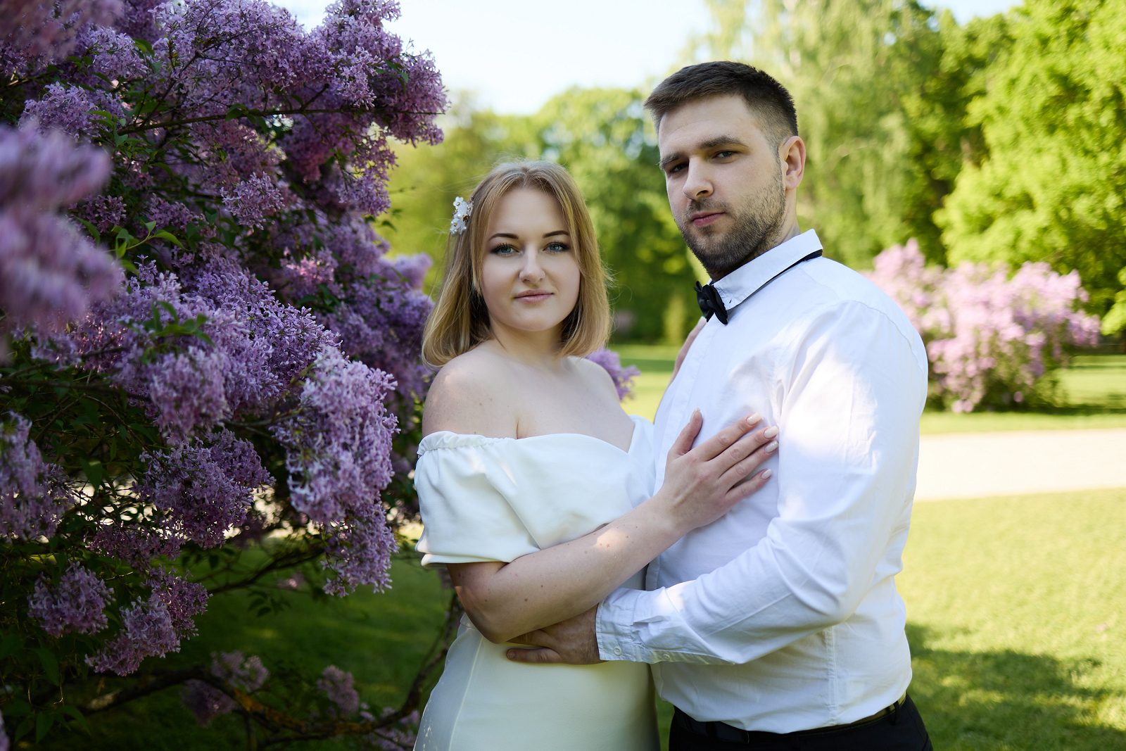 Pan młody obejmuje pannę młodą przy kwitnącym bzie w parku, a jej dłoń spoczywa na jego piersi.