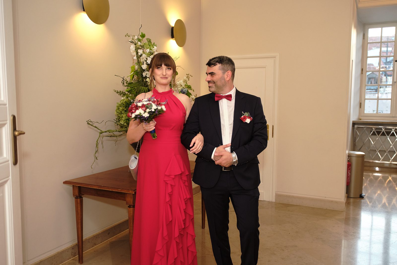 Nowożeńcy stoją obok siebie w sali ceremonii; panna młoda trzyma bukiet.