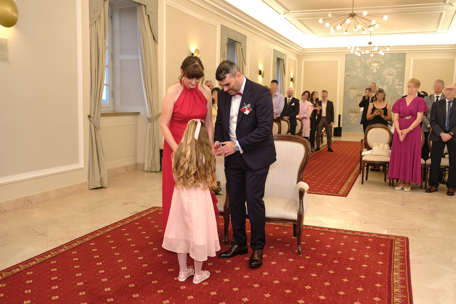 Panna młoda w czerwonej sukience pochyla się, by przywitać dziewczynkę w różowej.